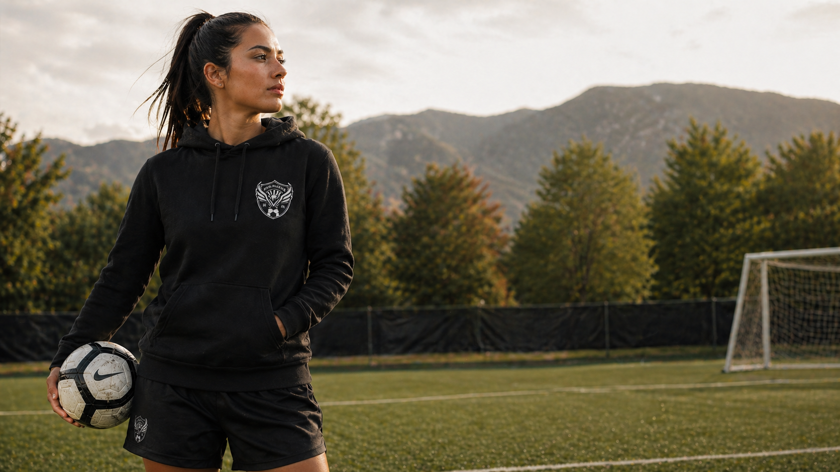Woman in black sports hoodie and shorts holding a soccer ball on a field with mountains in the background